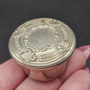 May include: A silver-toned, round container with a decorative lid. The lid features a central wreath design surrounded by floral and geometric patterns. The container is held in a hand against a dark background.