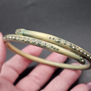 Pode incluir: Uma pulseira rígida de estilo bypass com um acabamento liso em verde-oliva pálido com efeito degradê. A pulseira é acentuada com uma única fileira de pequenos strass redondos e transparentes incrustados na superfície ao longo da banda.