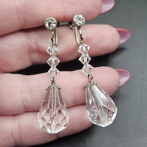 Può includere: Un paio di orecchini pendenti vintage in cristallo trasparente. Ogni orecchino presenta uno strass rotondo e trasparente nella parte superiore, seguito da tre perline di cristallo trasparente e un grande pendente di cristallo sfaccettato a forma di goccia.