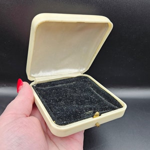 Könnte beinhalten: Eine weiße, rechteckige Schmuckschatulle mit schwarzem Samt-Innenfutter. Die Schachtel hat einen goldenen Verschluss.