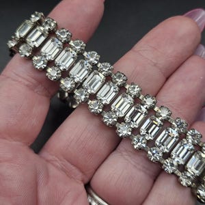 May include: A silver-toned bracelet with clear, rectangular and round rhinestones. The bracelet is composed of three rows of rectangular stones, with round stones between each row. The bracelet is held in a hand.