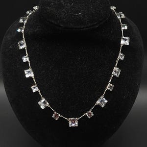 May include: A silver-toned necklace featuring a series of clear, square-cut faceted stones linked together in a single strand. The stones are set in metal frames and connected by thin, flexible metal bars, creating a uniform, repeating pattern.