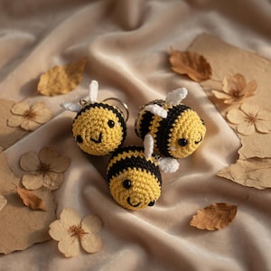 May include: Three handmade crochet bee keychains. Each bee features a yellow body with black stripes, white wings, and a smiling face. The keychains are set on a beige fabric background with dried leaves and flowers.