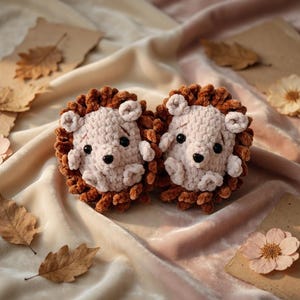 May include: Two handmade crochet lion toys with light pink faces, brown manes, and black eyes. The toys are placed on a cream-colored fabric with dried leaves and flowers scattered around. The overall aesthetic is soft and whimsical.