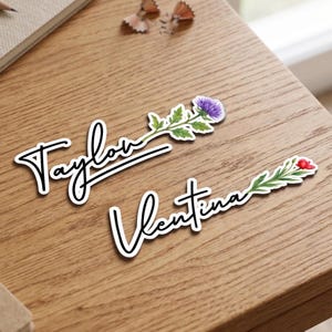 Può includere: Due adesivi personalizzati con i nomi "Taylor" e "Valentina", in corsivo con contorno bianco. L'adesivo "Taylor" ha un fiore di cardo viola e l'adesivo "Valentina" ha un fiore rosso.