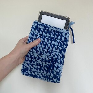 May include: A rectangular crochet sleeve made from thick, dark blue and light blue fabric yarn. The sleeve features a textured, interlocking stitch pattern and contains a black electronic tablet with a clear protective case inside the pouch.