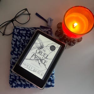 May include: A black e-reader displaying the book cover for The Cruel Prince by Holly Black, resting on a blue and light blue crocheted sleeve. Nearby are black-rimmed eyeglasses and a lit orange candle in a glass holder on a decorative coaster.