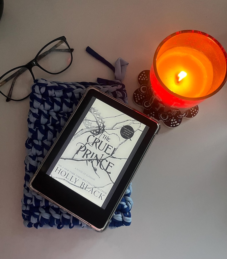 May include: A black e-reader displaying the book cover for The Cruel Prince by Holly Black, resting on a blue and light blue crocheted sleeve. Nearby are black-rimmed eyeglasses and a lit orange candle in a glass holder on a decorative coaster.