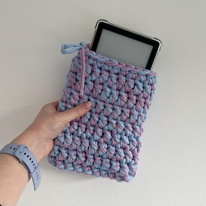May include: A rectangular crochet sleeve made from thick, light blue and lavender yarn, featuring a drawstring closure at the top. A black e-reader device with a clear protective case is partially inserted into the top opening of the sleeve.