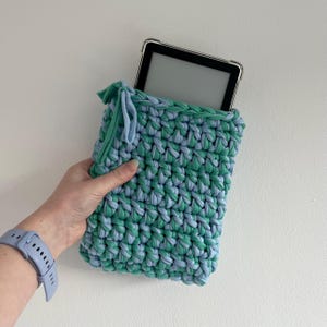 May include: A rectangular crochet sleeve made from thick, intertwined light blue and green fabric yarn. The sleeve features a drawstring closure at the top corner and contains a black electronic tablet with a clear protective case inside.