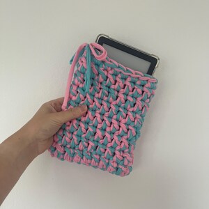 May include: A rectangular crochet sleeve made from thick, alternating pink and teal fabric yarn. The pouch features a drawstring closure at the top with matching pink and teal ties, designed to hold a black electronic tablet device securely.