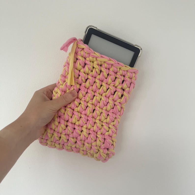 May include: A rectangular crochet sleeve made from thick, intertwined pink and yellow yarn. The sleeve features a small, light yellow fabric tag attached to the top left corner and contains a black electronic e-reader with a clear protective case.