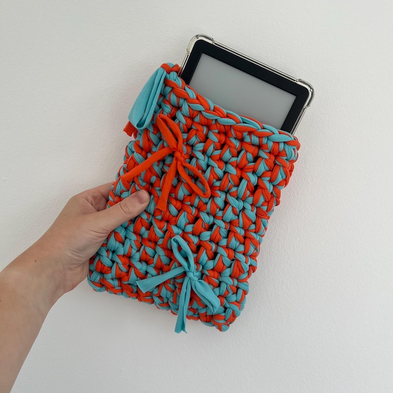 May include: A rectangular crochet sleeve made of thick, intertwined orange and light blue fabric strips. The sleeve features two decorative fabric bows, one orange and one light blue, and contains a black electronic e-reader device.