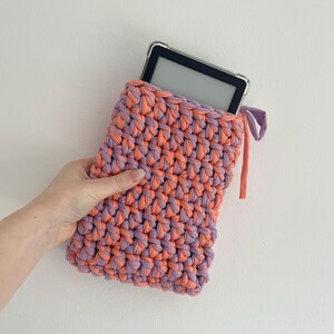 May include: A rectangular crochet sleeve made from thick, intertwined coral and lavender yarn. The sleeve holds a black electronic e-reader with a clear protective case. A small fabric tie in matching colors extends from the top right corner.