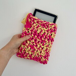 May include: A rectangular crochet sleeve made from thick, intertwined hot pink and pale yellow yarn. The sleeve features a small, solid pink fabric tag attached to the top left corner and contains a black electronic tablet with a clear case.