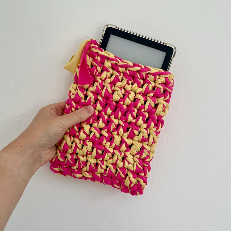 May include: A rectangular crochet sleeve made from thick, intertwined hot pink and pale yellow yarn. The sleeve features a small, solid pink fabric tag attached to the top left corner and contains a black electronic tablet with a clear case.