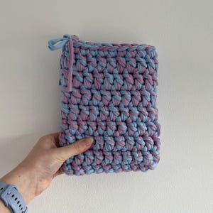 May include: A rectangular pouch crafted from thick, chunky yarn in alternating light blue and dusty purple colors. The crochet construction features a dense, textured stitch pattern with a small fabric tie closure at the top left corner.