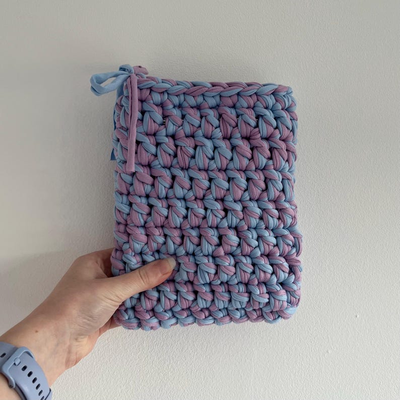 May include: A rectangular pouch crafted from thick, chunky yarn in alternating light blue and dusty purple colors. The crochet construction features a dense, textured stitch pattern with a small fabric tie closure at the top left corner.