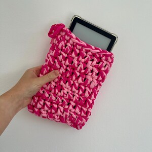 May include: A rectangular crochet sleeve made from thick, intertwined hot pink and light pink fabric yarn. The sleeve features a small pink ribbon tie at the top corner and contains a black electronic tablet with a clear protective case.