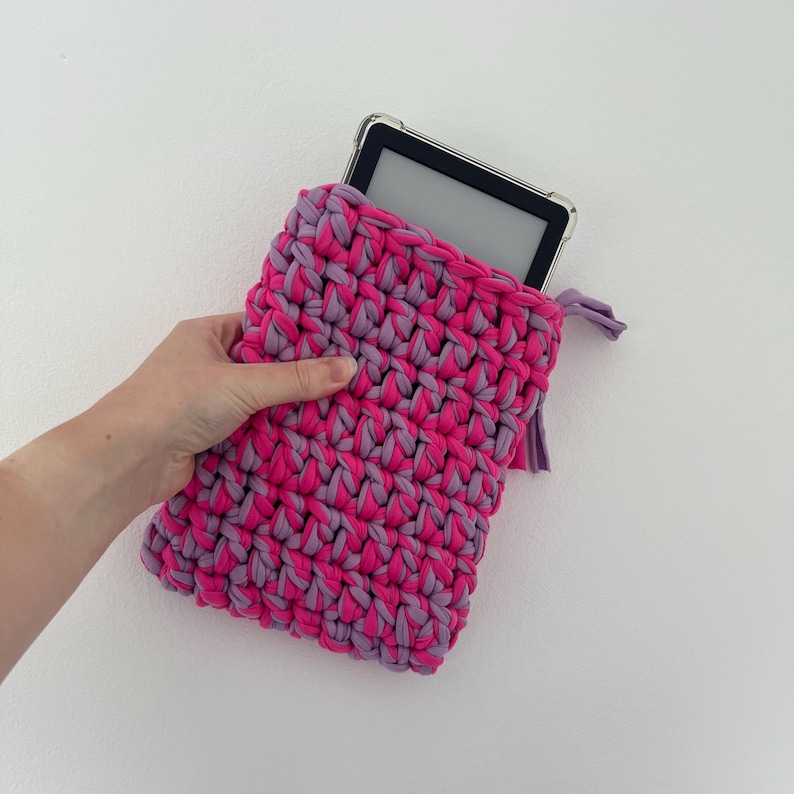 May include: A rectangular crochet sleeve made from thick, chunky yarn in alternating shades of bright fuchsia pink and light lavender purple. The sleeve contains a black electronic tablet with a clear protective case visible at the top opening.