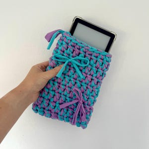 May include: A rectangular crochet sleeve made from thick, intertwined turquoise and purple fabric yarn. The sleeve features two decorative bows, one turquoise and one purple, and contains a black electronic tablet with a clear protective case.