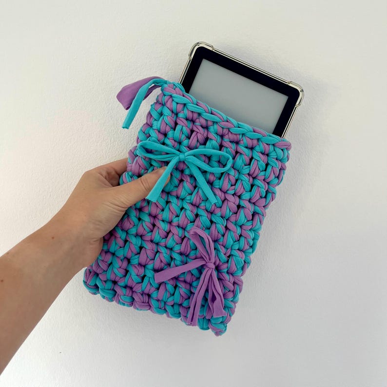 May include: A rectangular crochet sleeve made from thick, intertwined turquoise and purple fabric yarn. The sleeve features two decorative bows, one turquoise and one purple, and contains a black electronic tablet with a clear protective case.