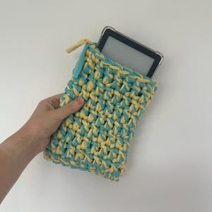 May include: A rectangular crochet sleeve made from thick, intertwined light blue and pale yellow yarn. The sleeve features a zipper closure at the top and contains a black electronic e-reader device with a clear protective case inside.
