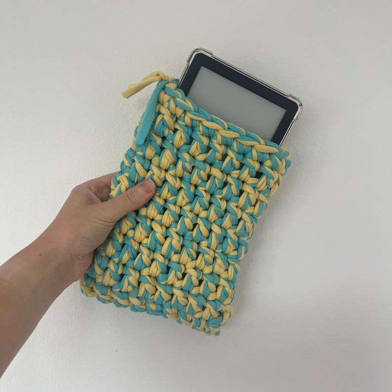 May include: A rectangular crochet sleeve made from thick, intertwined light blue and pale yellow yarn. The sleeve features a zipper closure at the top and contains a black electronic e-reader device with a clear protective case inside.