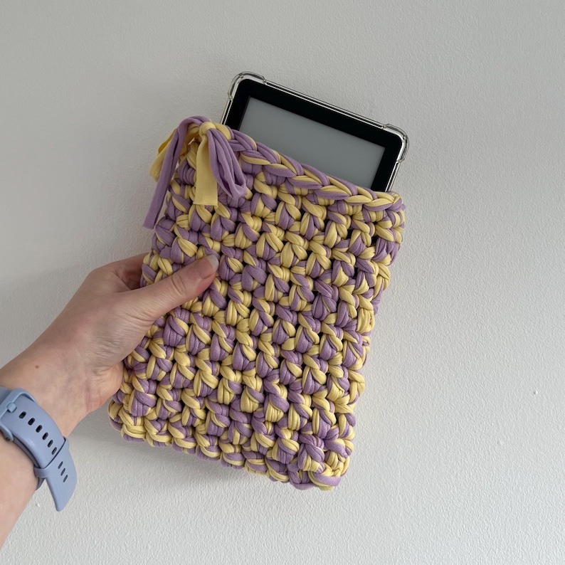 May include: A rectangular crochet sleeve made from thick, intertwined lavender and pale yellow yarn. The sleeve features a drawstring closure with matching fabric ties at the top corner and contains a black e-reader with a clear protective case.