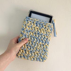 May include: A rectangular crochet sleeve made from thick, light blue and pale yellow fabric yarn. The sleeve features a textured, chunky knit pattern and contains a black e-reader device with a clear protective case visible at the top opening.