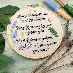 May include: A white embroidery hoop with a beige fabric background. The fabric has black cursive text that reads "Always throw salt over your left shoulder. Keep Rosemary by your garden gate. Plant Lavender for luck. And fall in love whenever you can." There are embroidered lavender flowers, rosemary sprigs, and gold stars.