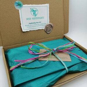 May include: A brown cardboard box with a teal blue tissue paper inside. The tissue paper is tied with a pink, green, and blue ribbon. The box has a sticker on the top that says "Heidi Hummingbird Daydreaming Since 1985 Thank you so much for supporting a teeny-weeny local business!"