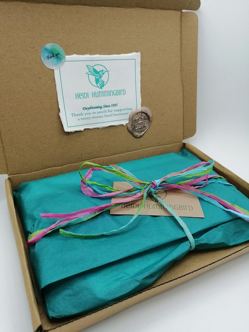 May include: A brown cardboard box with a teal blue tissue paper inside. The tissue paper is tied with a pink, green, and blue ribbon. The box has a sticker on the top that says "Heidi Hummingbird Daydreaming Since 1985 Thank you so much for supporting a teeny-weeny local business!"