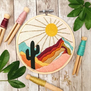 May include: A close-up of a completed embroidery hoop featuring a desert landscape scene. The embroidery depicts a cactus, mountains, and a sun with rays. The hoop is surrounded by embroidery floss and wooden clothes pegs.