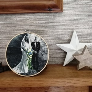 May include: A black and white photo of a couple in wedding attire, framed in a round wooden hoop. The bride is wearing a white dress and the groom is wearing a suit and tie. They are standing in front of a stone wall.