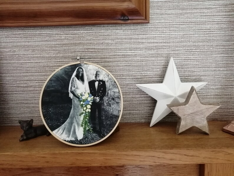 May include: A black and white photo of a couple in wedding attire, framed in a round wooden hoop. The bride is wearing a white dress and the groom is wearing a suit and tie. They are standing in front of a stone wall.