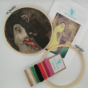 May include: A vintage photo of a couple on their wedding day, framed in a wooden embroidery hoop. The photo is in black and white and shows the couple standing in front of a building. The bride is wearing a white dress and veil, and the groom is wearing a suit. The photo is surrounded by a variety of colored embroidery floss.