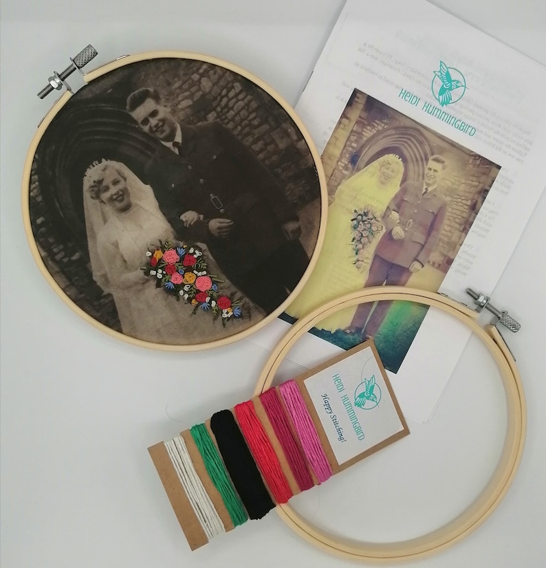 May include: A vintage photo of a couple on their wedding day, framed in a wooden embroidery hoop. The photo is in black and white and shows the couple standing in front of a building. The bride is wearing a white dress and veil, and the groom is wearing a suit. The photo is surrounded by a variety of colored embroidery floss.
