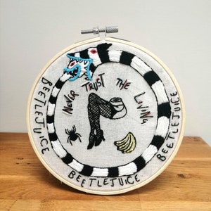 May include: Embroidered hoop art featuring a black and white striped design with the words "Beetlejuice" and "Never Trust The Living". The design also includes a cartoon monster with a blue tongue, a woman's legs in fishnet stockings, a spider, and bananas.