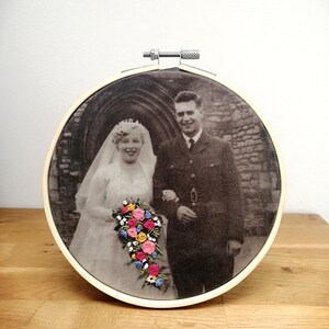 May include: A black and white wedding photo of a couple in front of a stone archway. The bride is wearing a white dress and veil, and the groom is wearing a military uniform. The photo is embroidered with a bouquet of colorful flowers.
