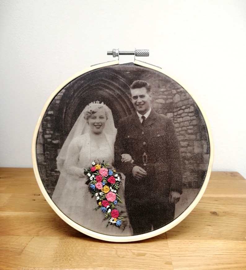 May include: A black and white wedding photo of a couple in front of a stone archway. The bride is wearing a white dress and veil, and the groom is wearing a military uniform. The photo is embroidered with a bouquet of colorful flowers.