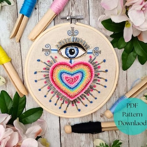 May include: A colorful embroidery hoop featuring a heart and eye design. The heart is filled with rainbow colors, and the eye is blue. The embroidery is set against a beige fabric. Wooden thread holders with blue, pink, and yellow thread are also visible.