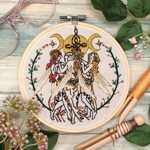 May include: Embroidered hoop art featuring three figures with wings, surrounded by floral vines and a triple moon symbol. The figures are depicted in shades of pink, yellow, and green against a white background.