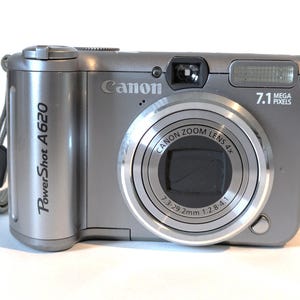 May include: A silver Canon PowerShot A620 digital camera with a zoom lens and 7.1 megapixels. The camera features a built-in flash and a wrist strap. The lens has a focal length of 7.3-29.2mm.