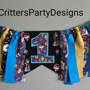 May include: A colorful fabric banner with a black felt pennant featuring the number "1" and characters from the movie "Coco". The banner is decorated with blue, yellow, and black fabric strips.