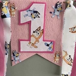 May include: Pink felt banner with a large number 1 and cartoon character cutouts. The banner has a pink border and is decorated with cartoon characters. The banner is for a first birthday celebration.