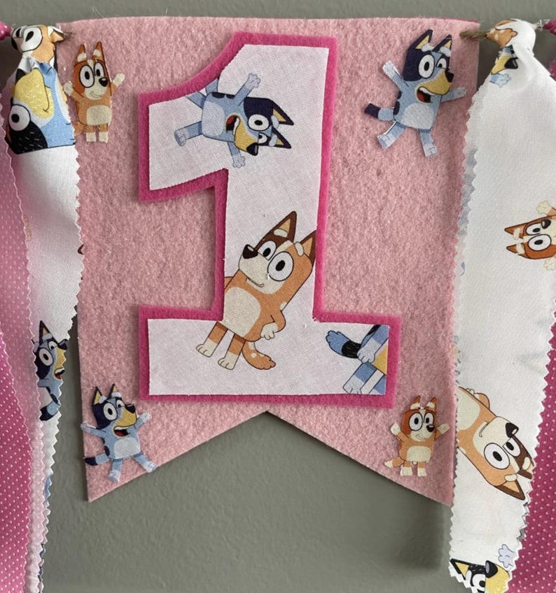 May include: Pink felt banner with a large number 1 and cartoon character cutouts. The banner has a pink border and is decorated with cartoon characters. The banner is for a first birthday celebration.