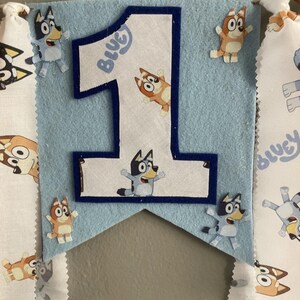May include: A light blue felt banner with a large number one, featuring the word "Bluey". The banner is decorated with cartoon character cutouts. Additional fabric banners with cartoon characters are attached to the sides.