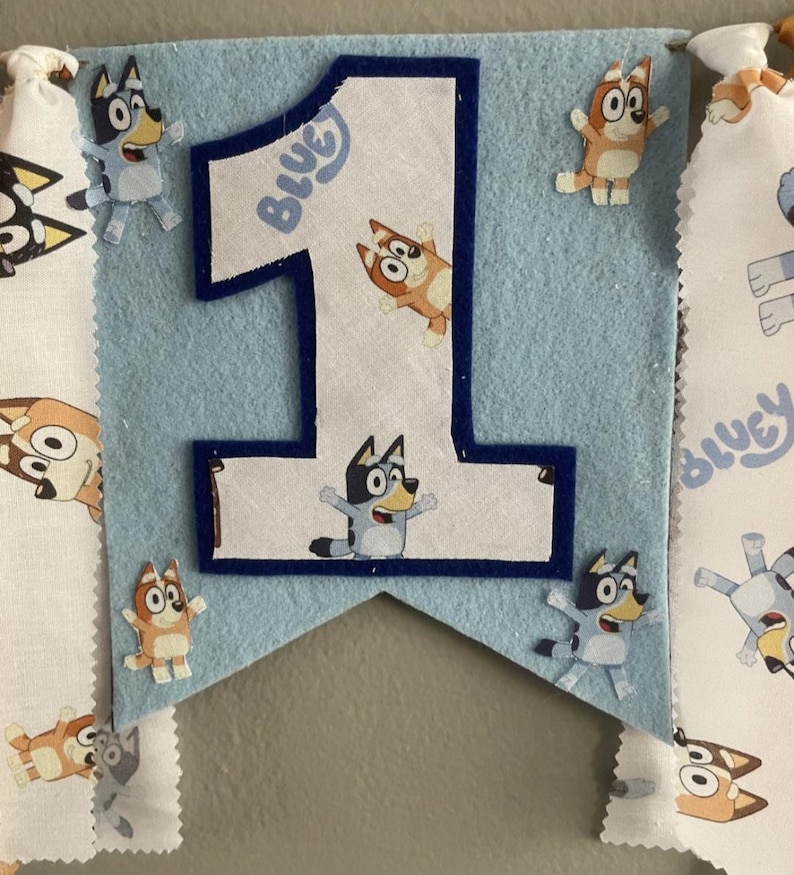 May include: A light blue felt banner with a large number one, featuring the word "Bluey". The banner is decorated with cartoon character cutouts. Additional fabric banners with cartoon characters are attached to the sides.