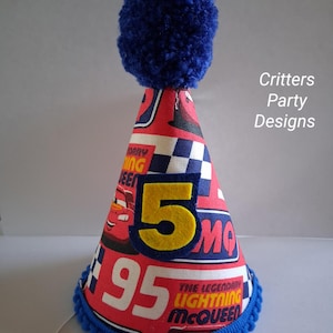 May include: A red and white party hat with a blue pom-pom on top. The hat features the number "5" in yellow, the number "95" in white, and images of Lightning McQueen. The bottom edge is trimmed with blue pom-poms. The text "Critters Party Designs" is visible.